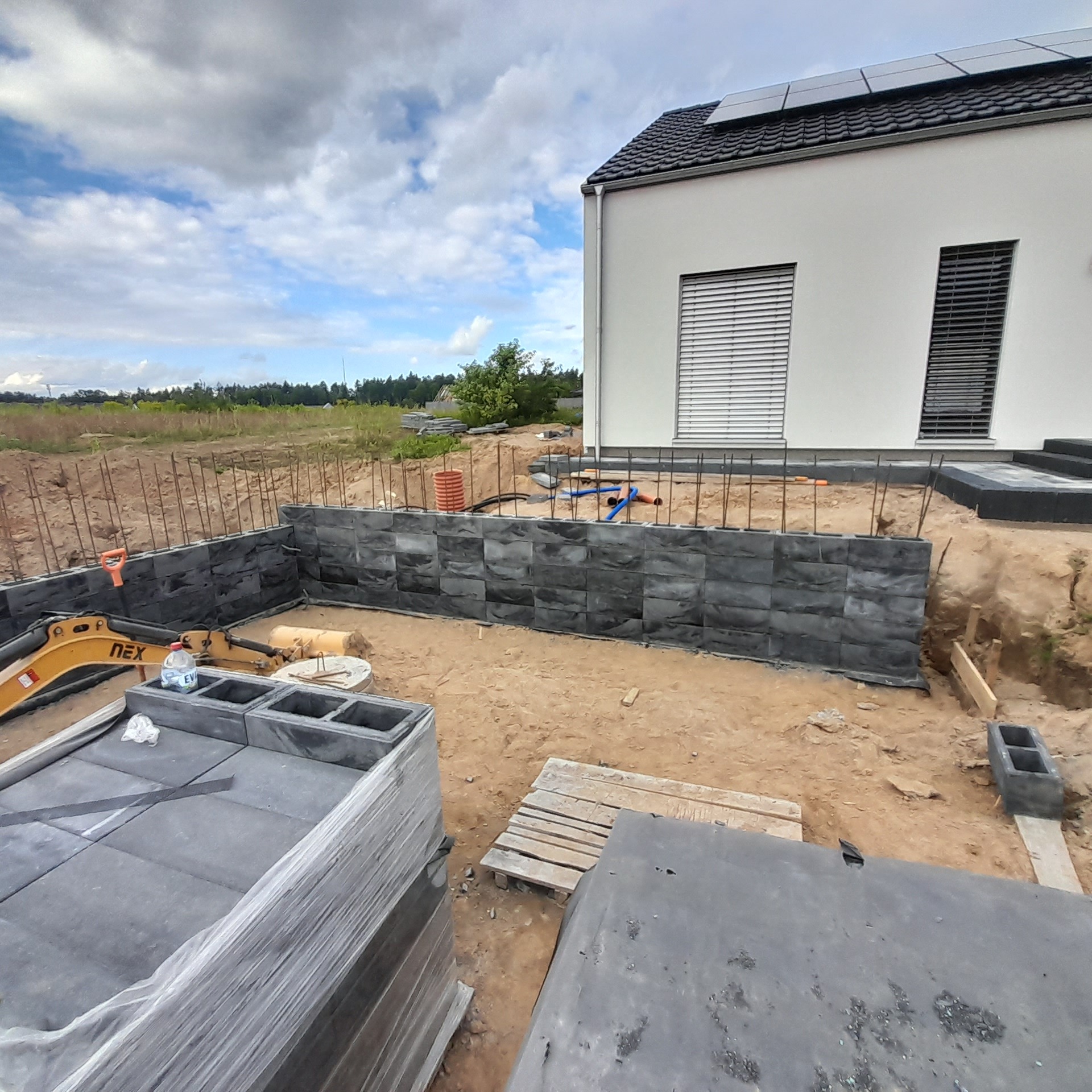 Budowa fundamentów z bloczków betonowych, widoczny wykop, zbrojenie pionowe, mini koparka i paleta z bloczkami, na drugim planie dom z panelami słonecznymi.