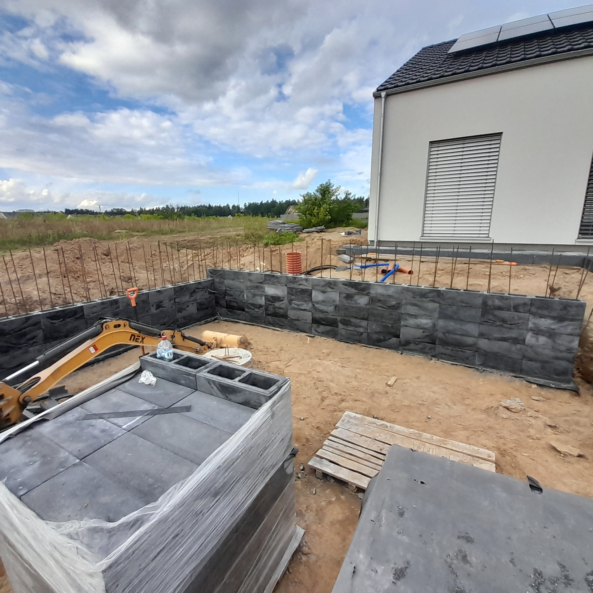 Budowa fundamentów z wykorzystaniem szarych bloczków, widoczny mały żółty koparka i zbrojenie, w tle dom z panelami słonecznymi.