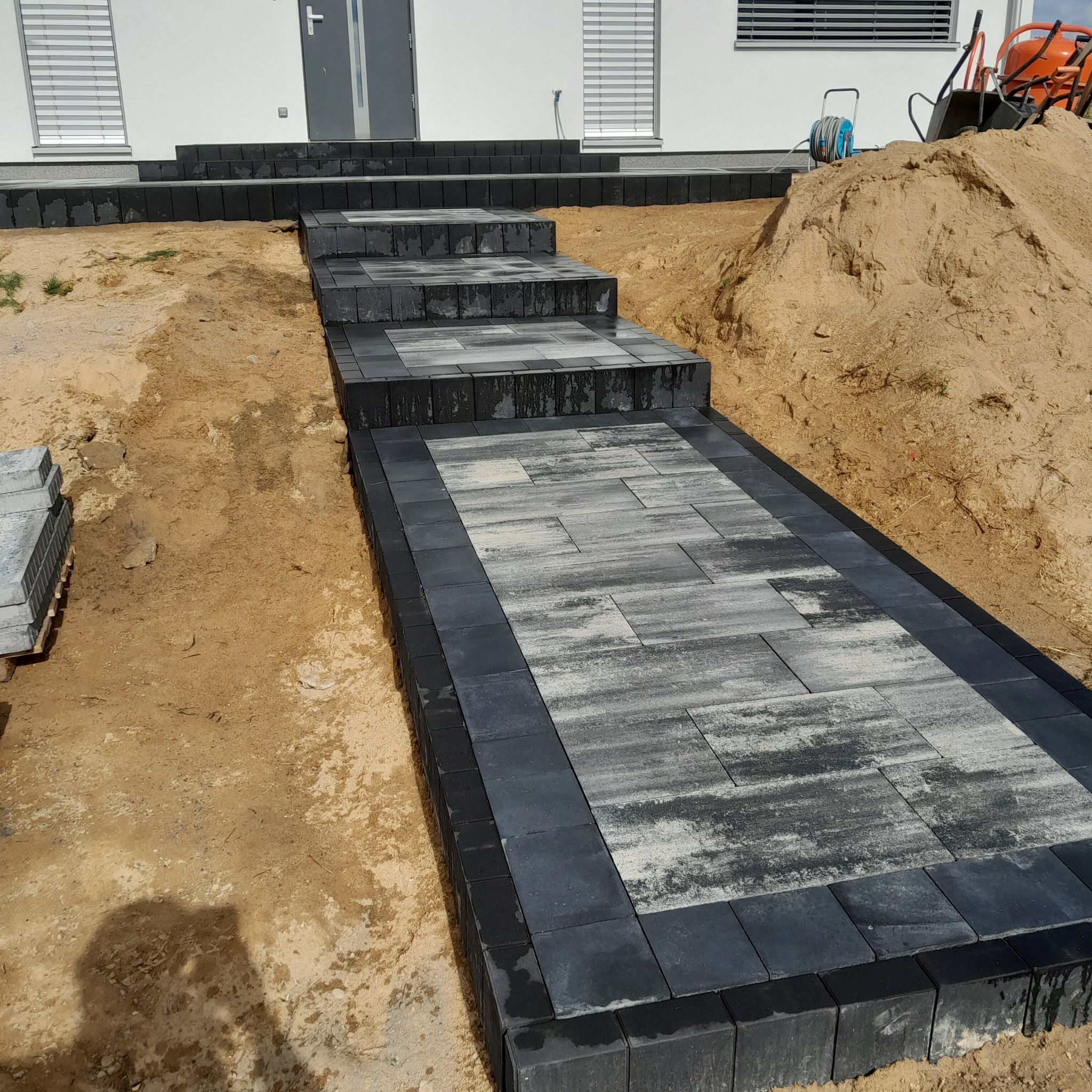 Schody i chodnik z kostki brukowej w odcieniach szarości, prowadzące do nowoczesnego domu z widoczną stertą piasku i narzędziami w tle.