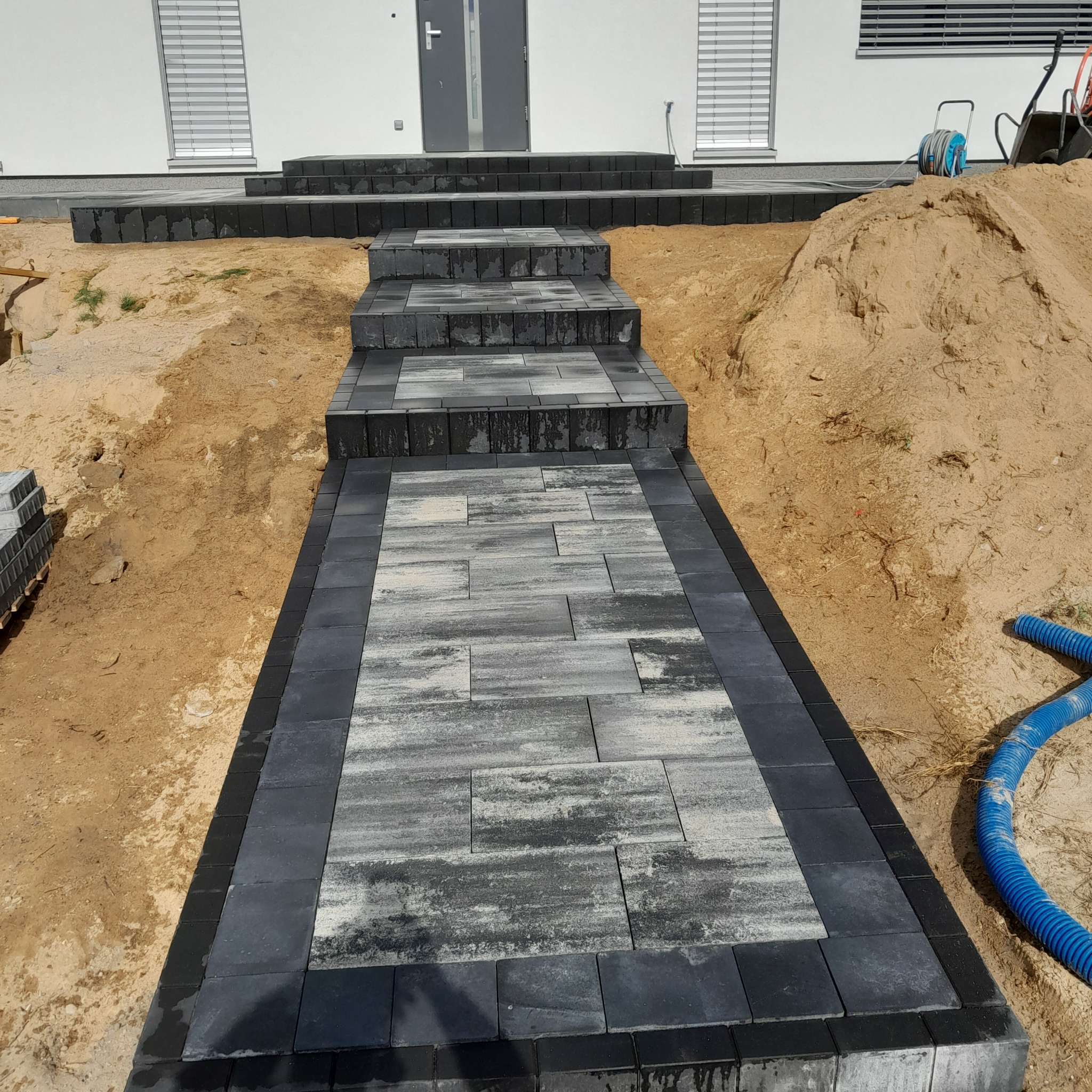 Schody i ścieżka z kostki brukowej w odcieniach szarości, prowadzące do domu z jasną elewacją i szarymi oknami, teren wokół w trakcie prac ziemnych.