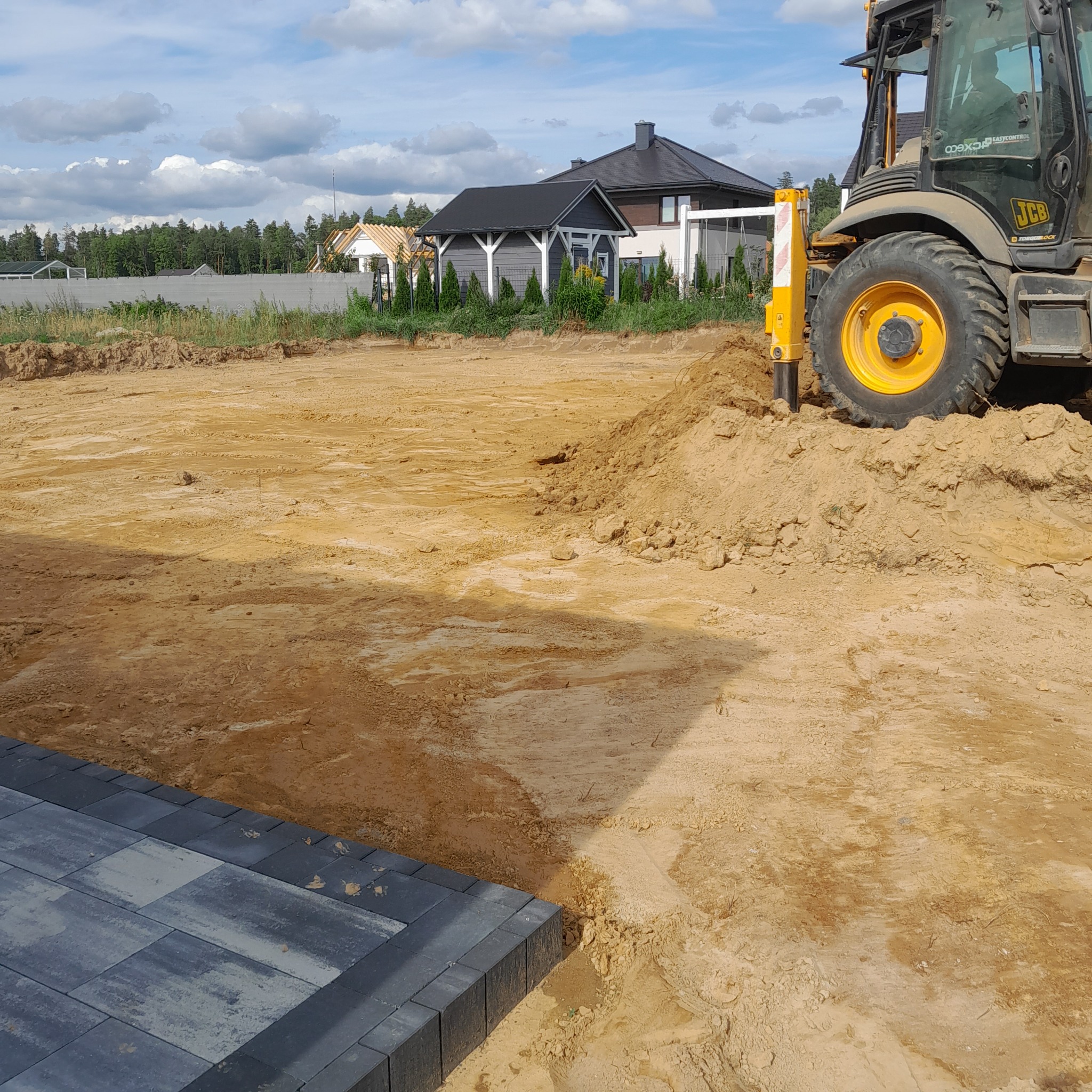Żółty traktor JCB ubija ziemię pod budowę, widoczne fragmenty ułożonej kostki brukowej, w tle domy jednorodzinne i las.