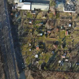 Widok z lotu ptaka na działki rekreacyjne z małymi domkami, ogrodami i ścieżkami, przylegające do linii kolejowej.