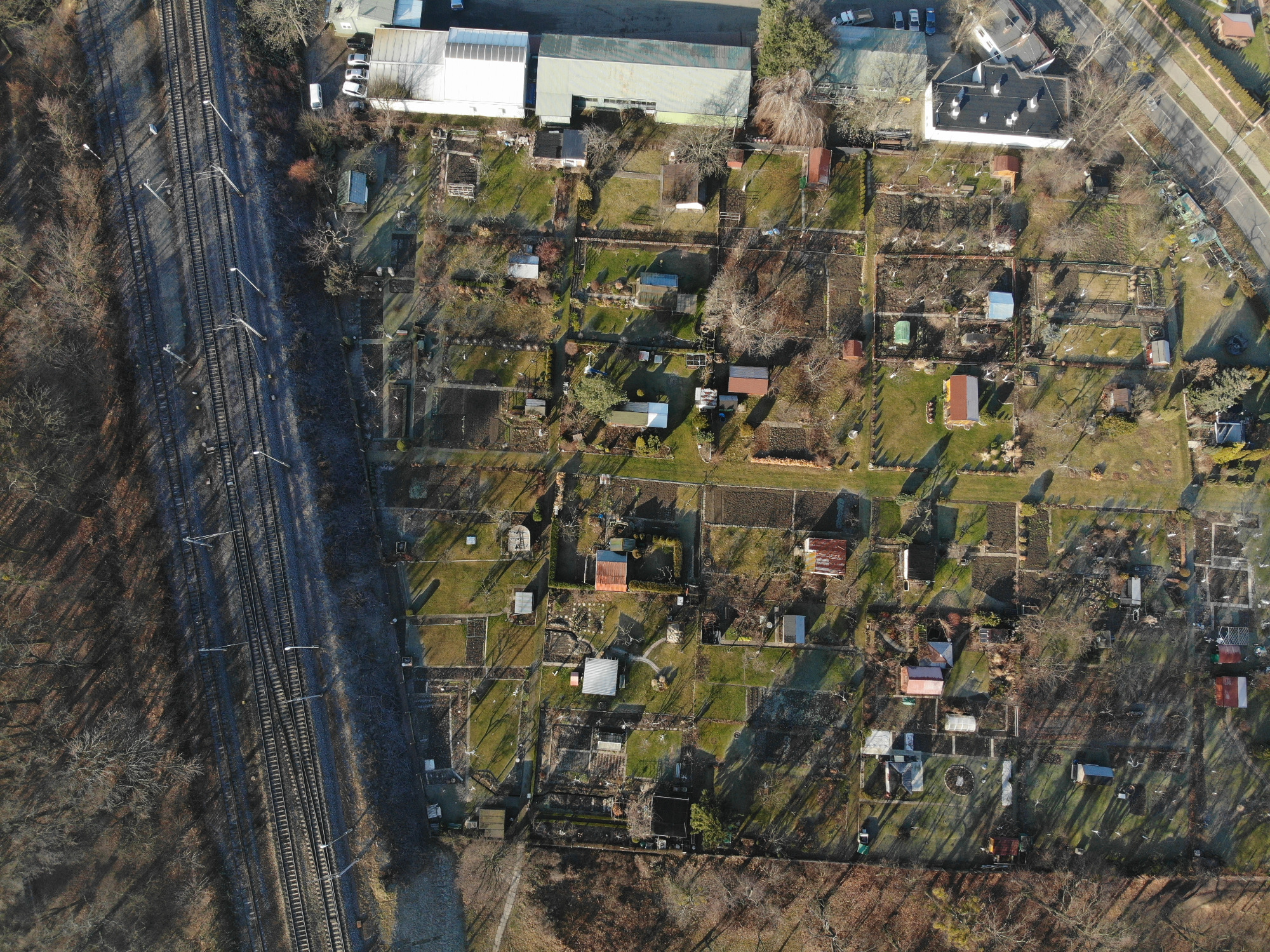 Widok z lotu ptaka na działki rekreacyjne z małymi domkami, ogrodami i ścieżkami, przylegające do linii kolejowej.