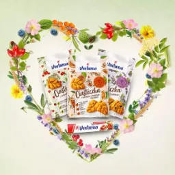 Trzy opakowania ciasteczek Verbena i batonik Verbena ułożone w kształt serca z polnych kwiatów, jagód i ziół na jasnym tle.