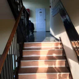 Klatka schodowa z drewnianymi schodami i metalową balustradą, na błyszczącej podłodze widać osobę w korytarzu przed białymi drzwiami z numerem 101.