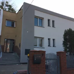 Dwukondygnacyjny dom w trakcie ocieplania elewacji, z częściowo ocieploną ścianą w kolorze beżowym i drugą częścią pomalowaną na biało-szaro, widoczne rusztowania z boku budynku, szare schody...