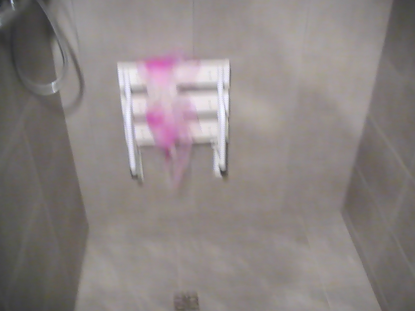 Niedokładne zdjęcie fragmentu łazienki z szarymi płytkami, widocznym prysznicem i składanym siedziskiem pod prysznicem, na którym wisi różowy ręcznik.