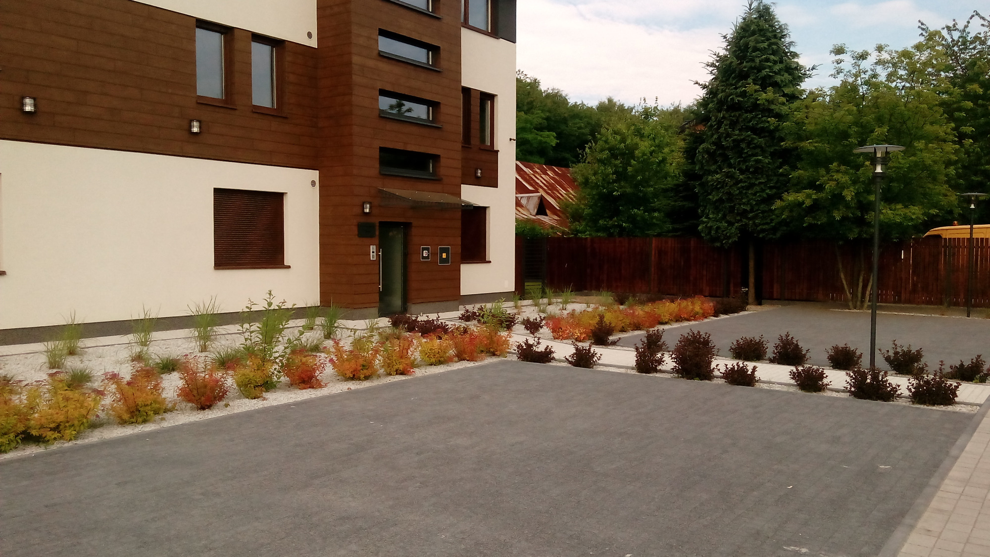 Nowoczesny budynek z brązową drewnianą elewacją i białymi ścianami, otoczony rabatami z ozdobnych krzewów i traw wysypanych białym kamieniem, na pierwszym planie szara kostka brukowa.
