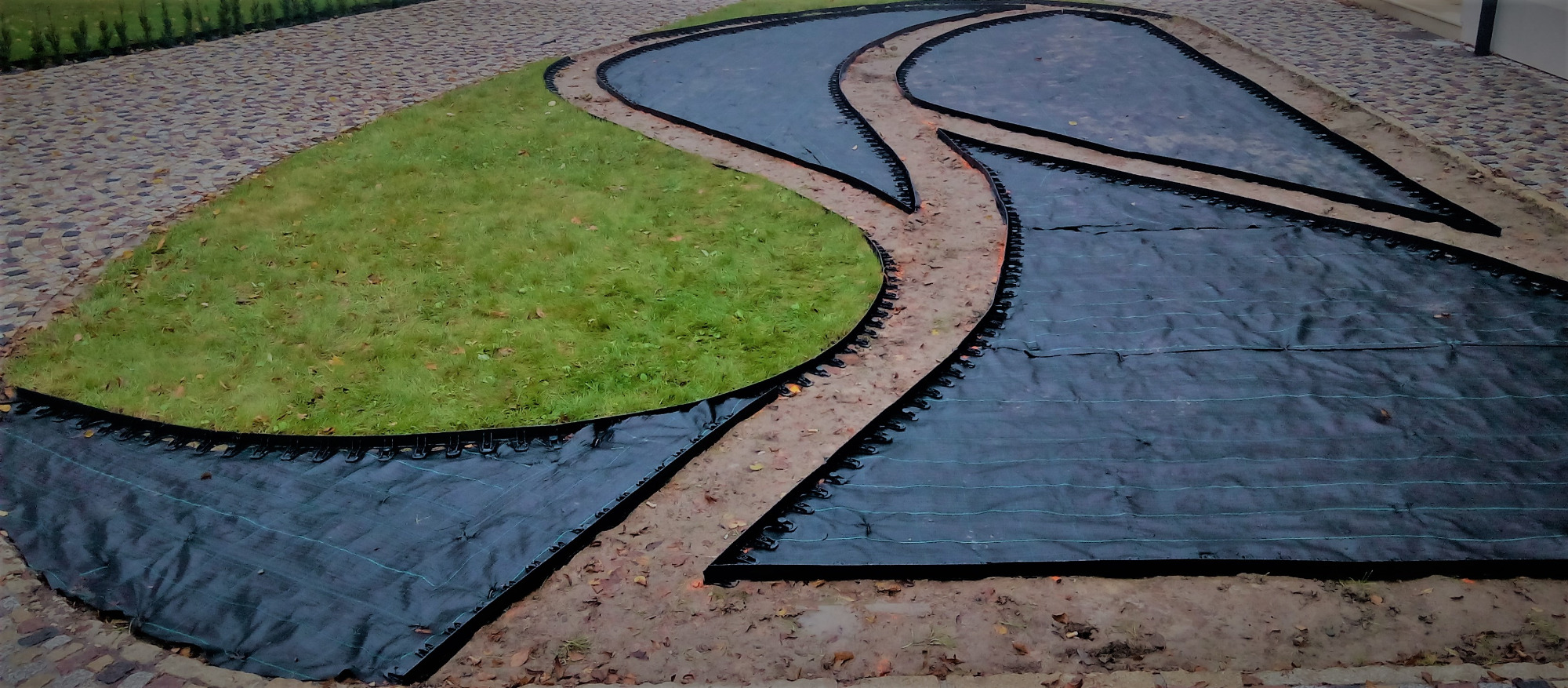 Przygotowanie terenu pod ogród: rozłożona czarna agrowłóknina z plastikową obwódką, wytyczająca ścieżki i rabaty na trawniku.