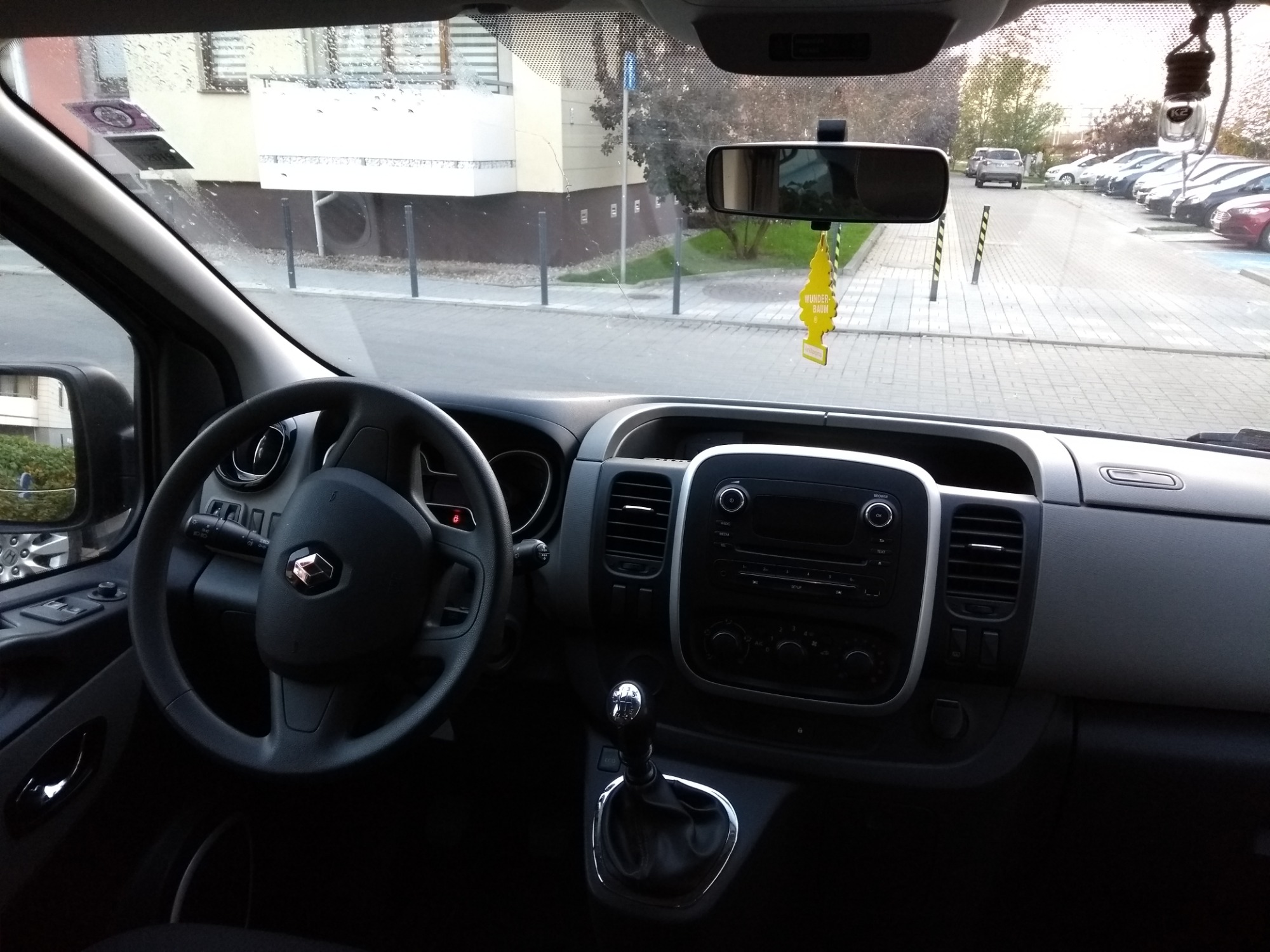 Wnętrze busa Renault z widocznym żółtym odświeżaczem powietrza Wunder-Baum wiszącym na lusterku wstecznym, na zewnątrz widoczny parking.