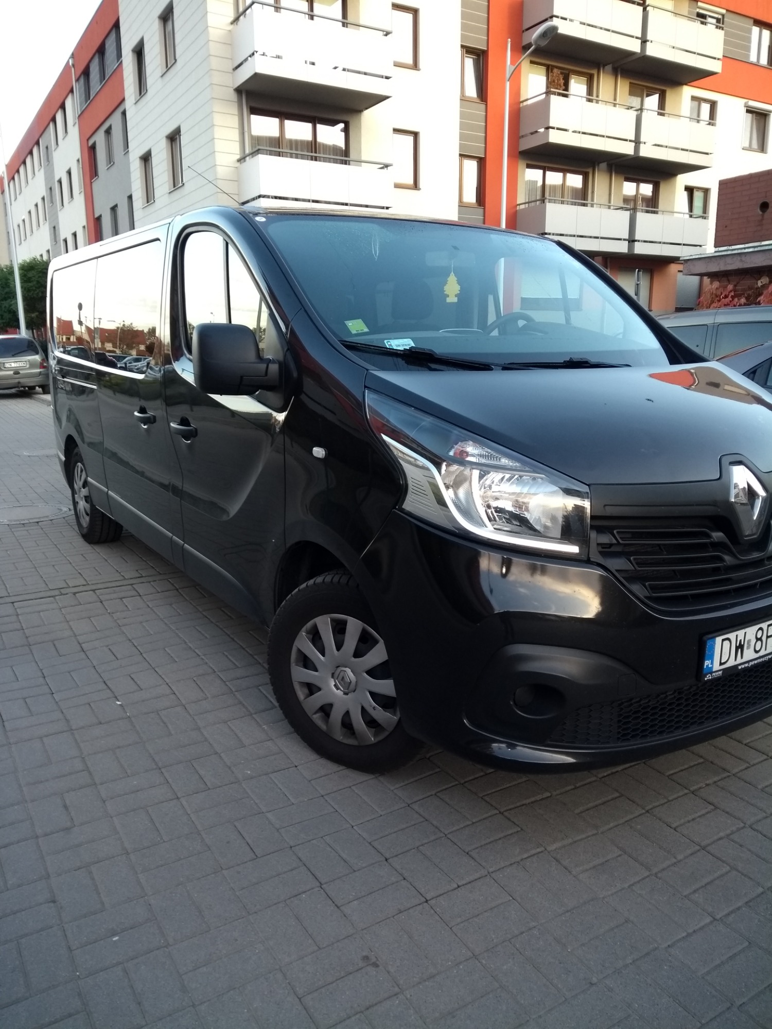 Czarny Renault Trafic zaparkowany na chodniku z kostki brukowej, widoczny budynek mieszkalny w tle, zawieszka zapachowa na lusterku.