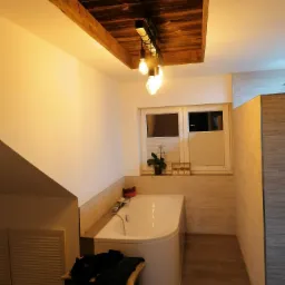 Nowoczesna łazienka z wanną, oświetleniem w drewnianej obudowie sufitowej i oknem z roletami plisowanymi, ubrania na krześle.