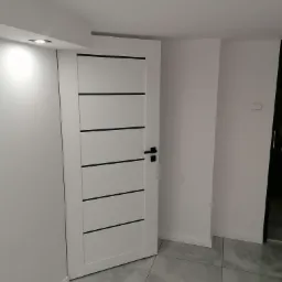 Nowoczesne, białe drzwi wewnętrzne z czarnymi, poziomymi akcentami w minimalistycznym wnętrzu z szarą, kwadratową podłogą z płytek i białymi ścianami.