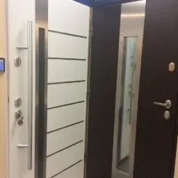 Wystawa dwóch modeli drzwi antywłamaniowych marki Gerda: białe z poziomymi, czarnymi paskami i srebrnym panelem oraz ciemnobrązowe z długim, pionowym, srebrnym panelem.