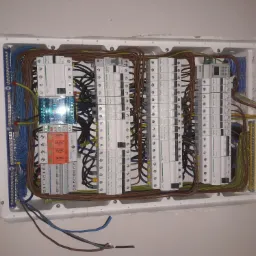 Rozdzielnica elektryczna w obudowie podtynkowej, widoczne bezpieczniki, wyłączniki różnicowoprądowe, listwy zaciskowe i przewody o różnych kolorach, zamontowana w ścianie z widocznymi...