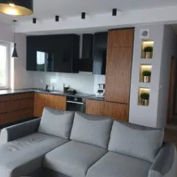 Nowoczesna kuchnia z drewnianymi i czarnymi szafkami, widoczna z perspektywy salonu z szarą sofą w Pyszącej. Na ścianie zegar i dekoracyjne wnęki z roślinami.