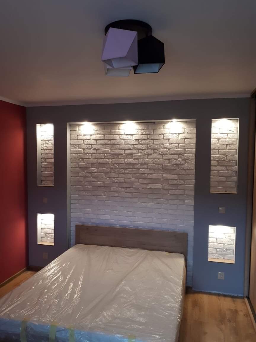 Pokój sypialny z łóżkiem przykrytym folią ochronną, ściana za wezgłowiem wykończona cegłą dekoracyjną z oświetleniem punktowym, nowoczesna lampa sufitowa.