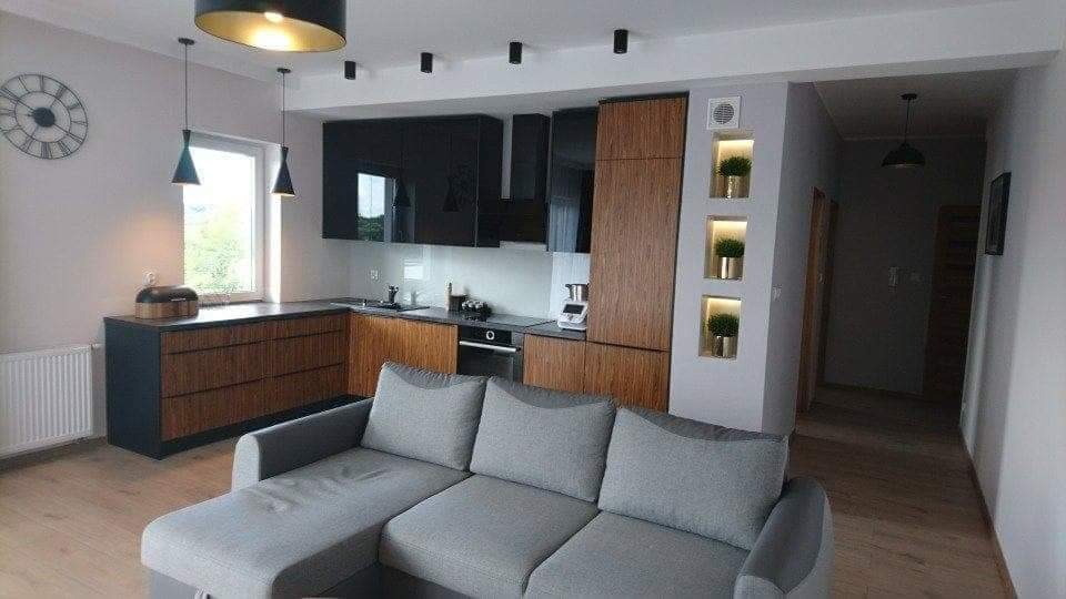 Nowoczesna kuchnia z drewnianymi i czarnymi szafkami, widoczna z perspektywy salonu z szarą sofą w Pyszącej. Na ścianie zegar i dekoracyjne wnęki z roślinami.