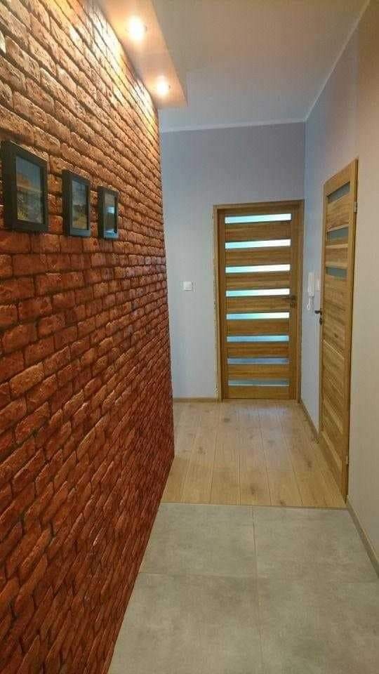 Korytarz po remoncie: ściana z cegły dekoracyjnej, drewniane drzwi z przeszkleniami, podłoga z płytek i paneli, oświetlenie punktowe.