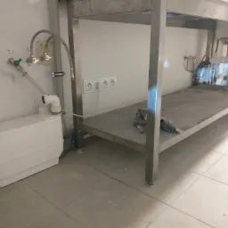 Biała pompa do ścieków zamontowana przy ścianie obok metalowego stołu z umywalką; widoczne rury i zawory doprowadzające wodę oraz gniazdka elektryczne na ścianie.