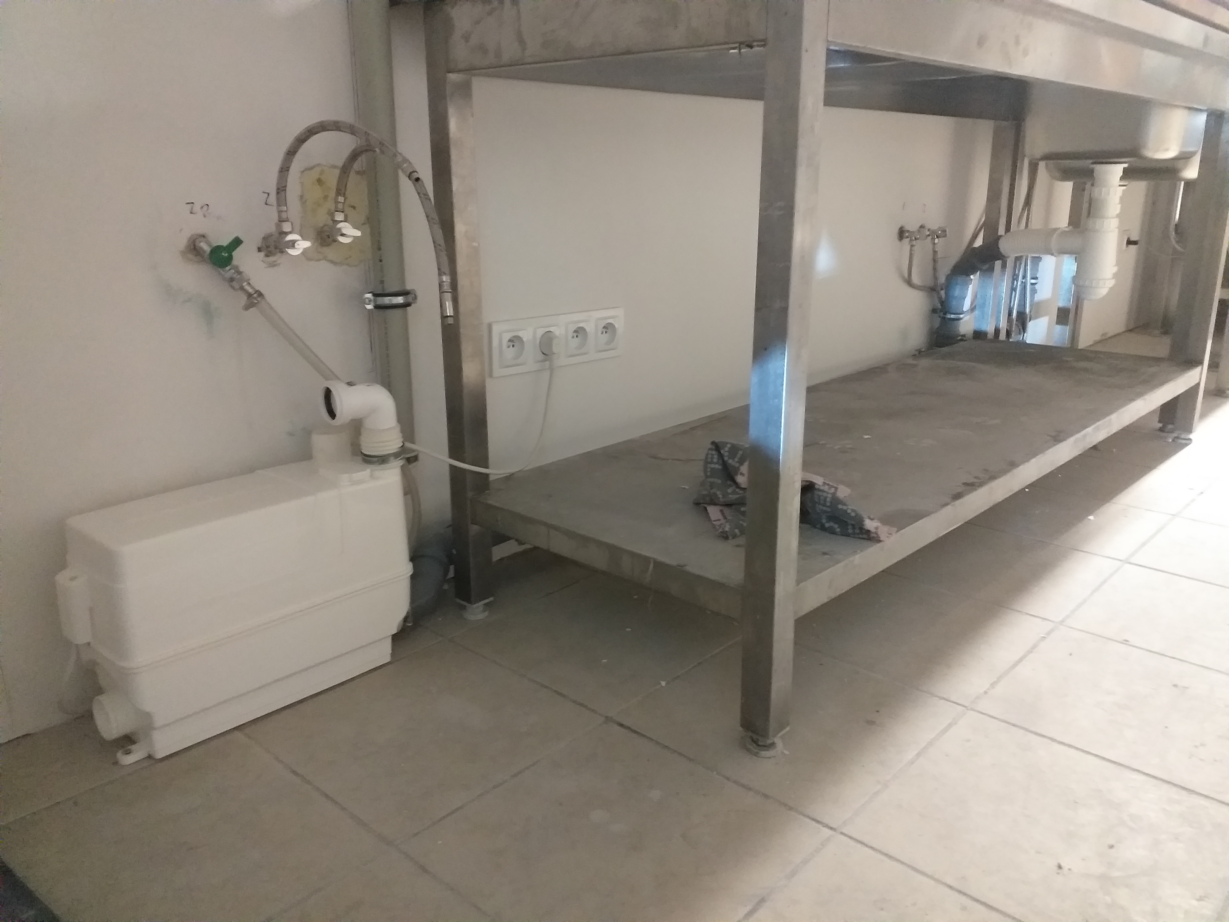 Biała pompa do ścieków zamontowana przy ścianie obok metalowego stołu z umywalką; widoczne rury i zawory doprowadzające wodę oraz gniazdka elektryczne na ścianie.