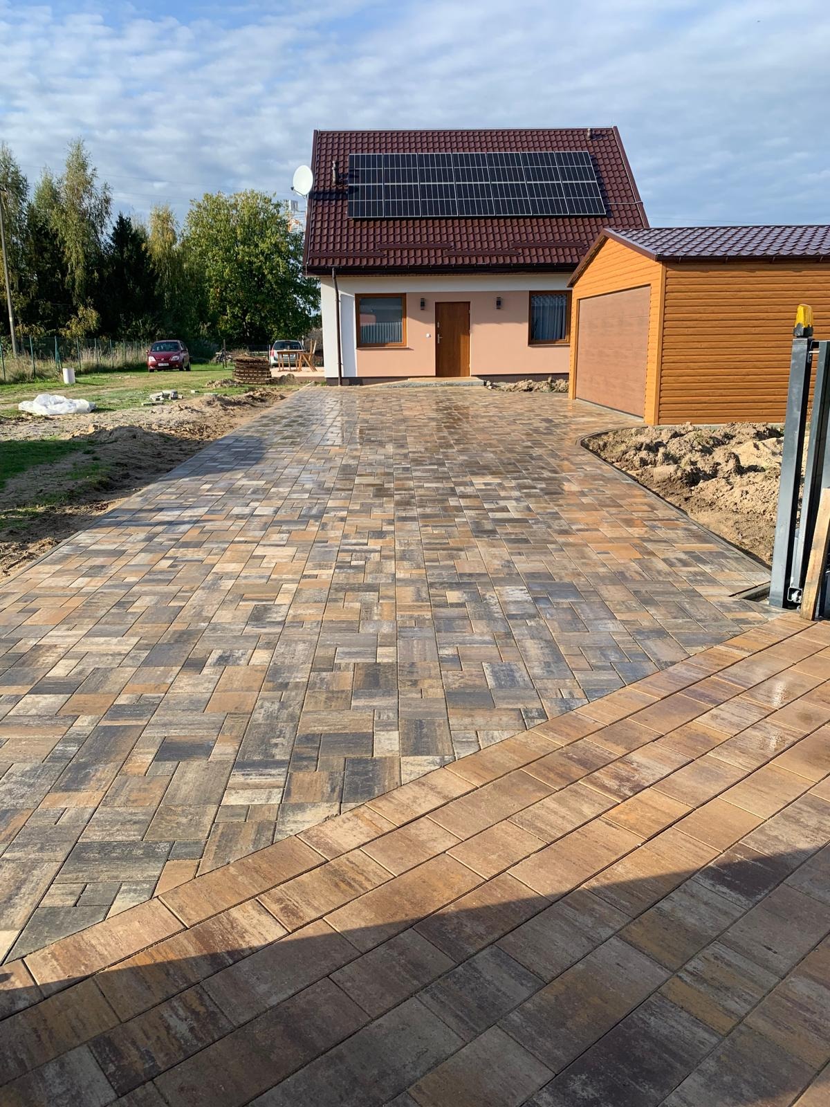 Nowoczesna, wielobarwna kostka brukowa na podjeździe domu z panelami słonecznymi. Widoczny garaż i fragment ogrodzenia.