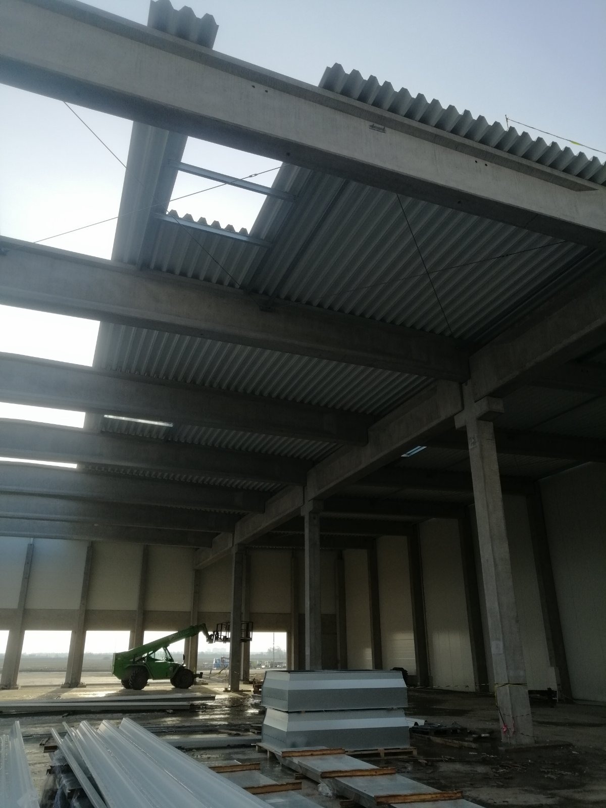 Wnętrze hali przemysłowej w trakcie budowy dachu z widocznymi betonowymi belkami, blachą trapezową i podnośnikiem teleskopowym w tle.