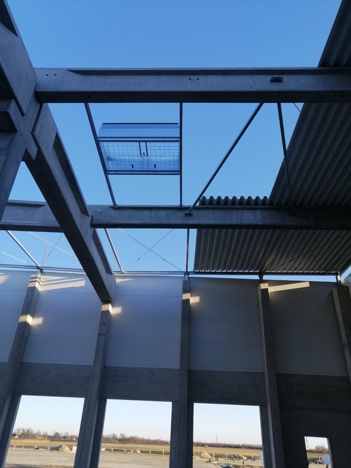 Wnętrze hali w trakcie budowy, widok z dołu na betonowe konstrukcje nośne, fragment dachu z blachy falistej i świetlik dachowy z kratą, błękitne niebo w tle.