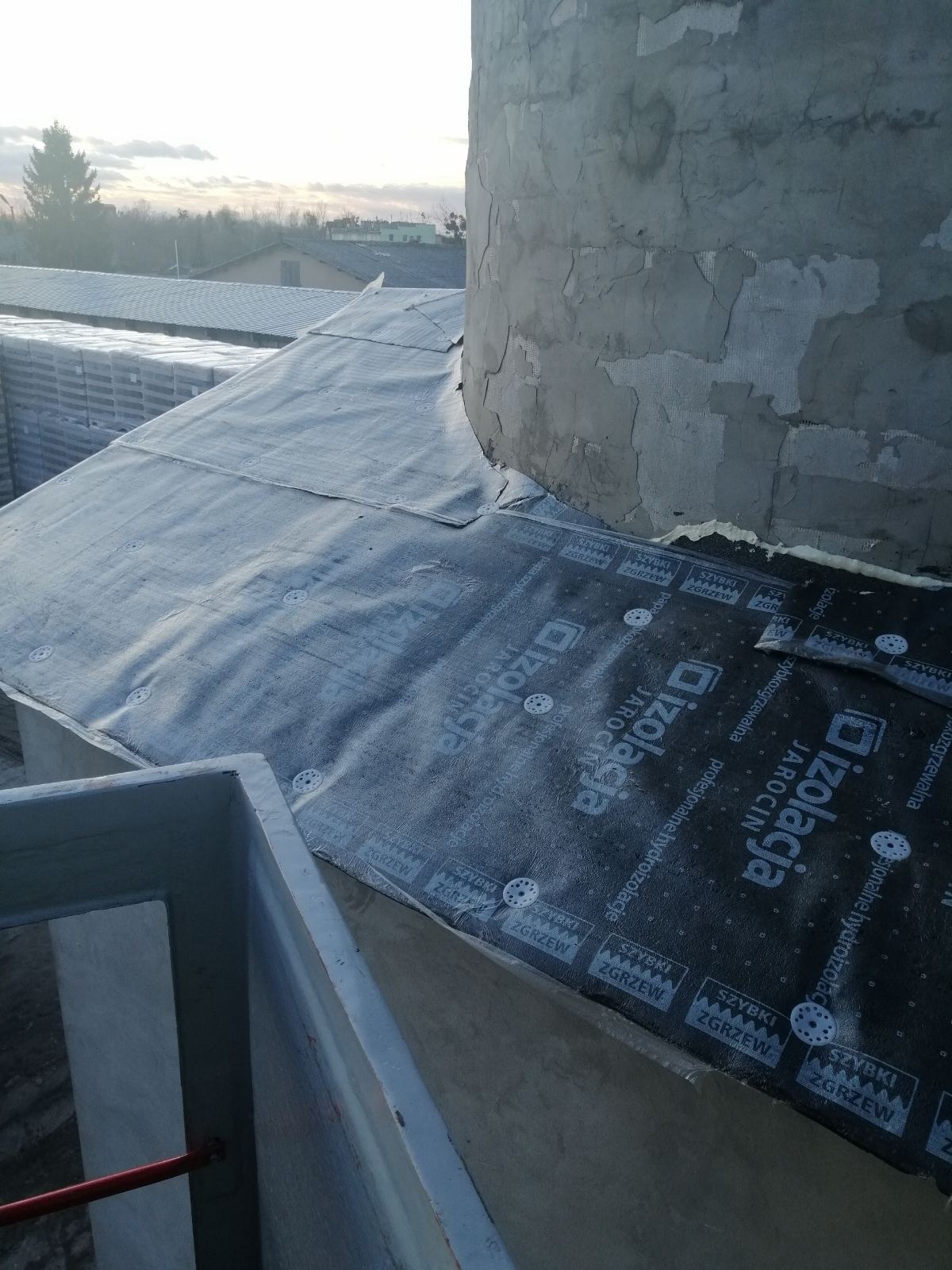Czarny, hydroizolacyjny materiał dachowy marki Izolacja Jarocin, przymocowany do dachu za pomocą białych kołków, przylegający do okrągłej, szarej konstrukcji komina z widocznymi ubytkami tynku.
