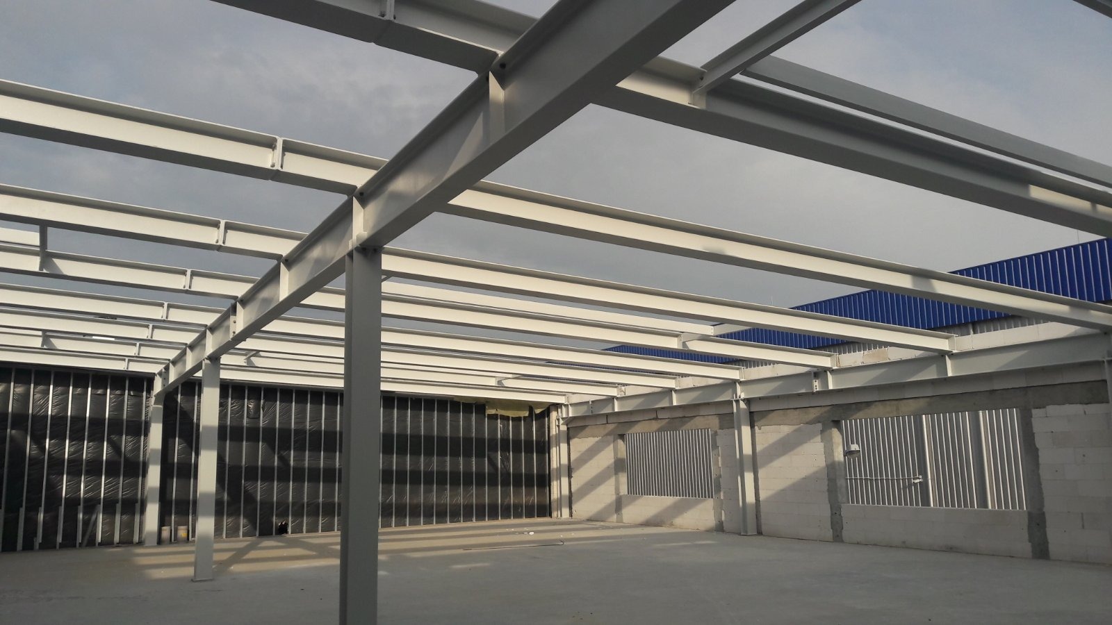 Konstrukcja stalowa dachu budynku przemysłowego, widok z wnętrza, szare niebo w tle, ściany z pustaków i blachy trapezowej.
