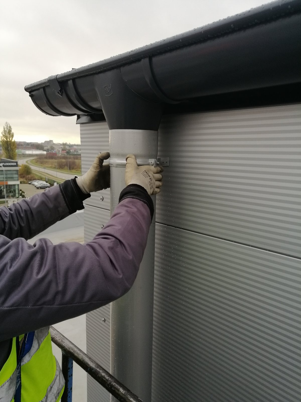 Montaż pionowej rury spustowej rynny na budynku o szarej elewacji panelowej, widoczne ręce w roboczych rękawicach mocujące obejmę, w tle fragment dachu i krajobraz miejski pod pochmurnym niebem.