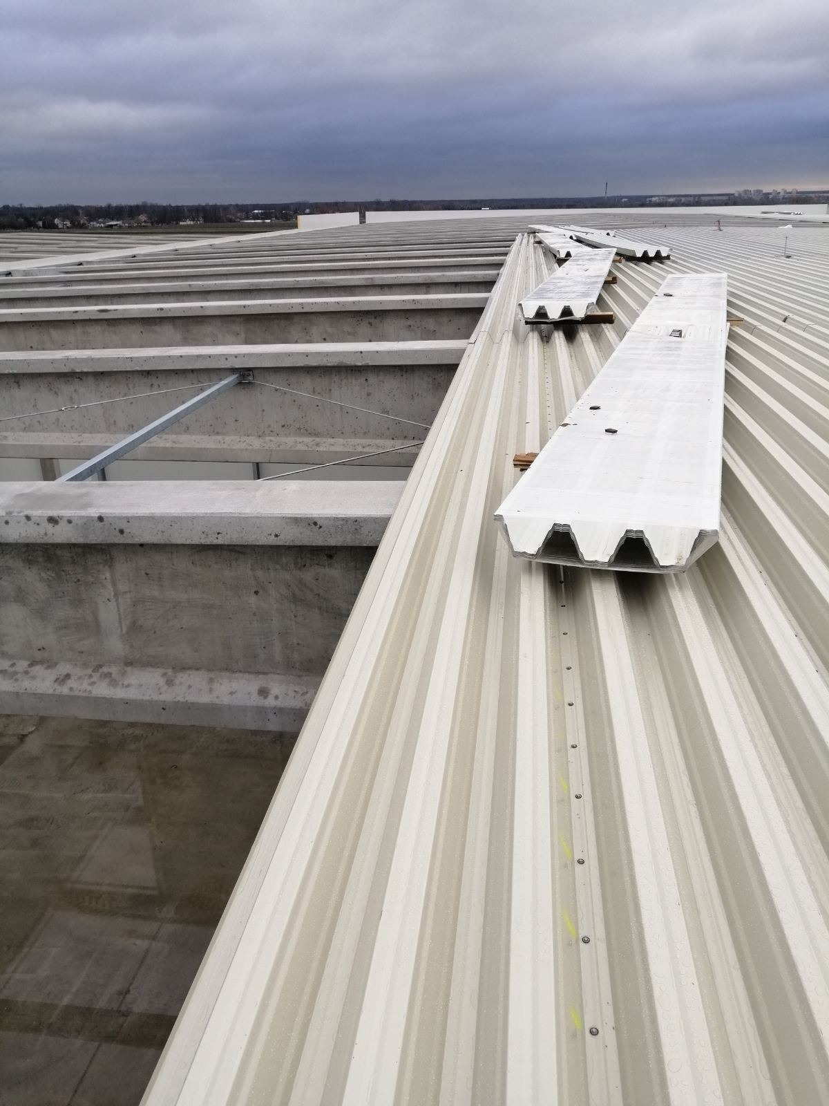 Montaż paneli dachowych z widocznymi betonowymi konstrukcjami nośnymi i pochmurnym niebem w tle.