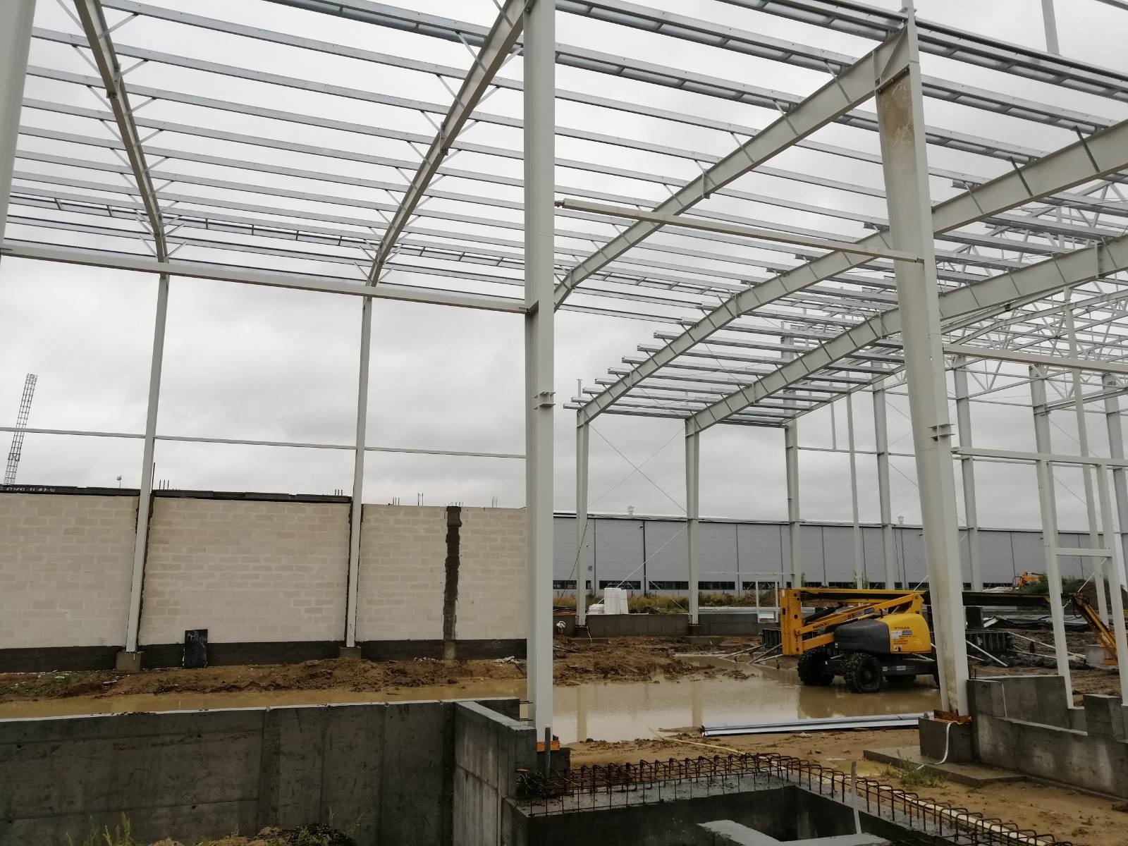 Konstrukcja stalowa dachu hali w trakcie budowy, widoczna żółta platforma robocza, ściany z bloczków betonowych, teren podmokły po deszczu.