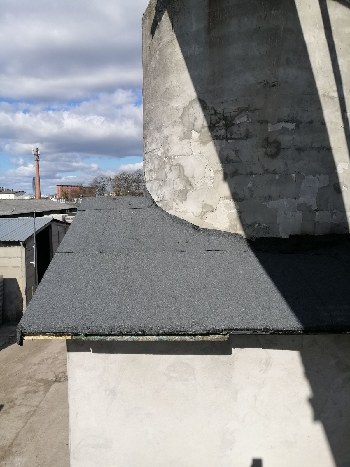 Płaski dach pokryty papą na tle betonowego silosu i zabudowań przemysłowych w oddali, widoczne nierówności i łączenia papy, cień na silosie.