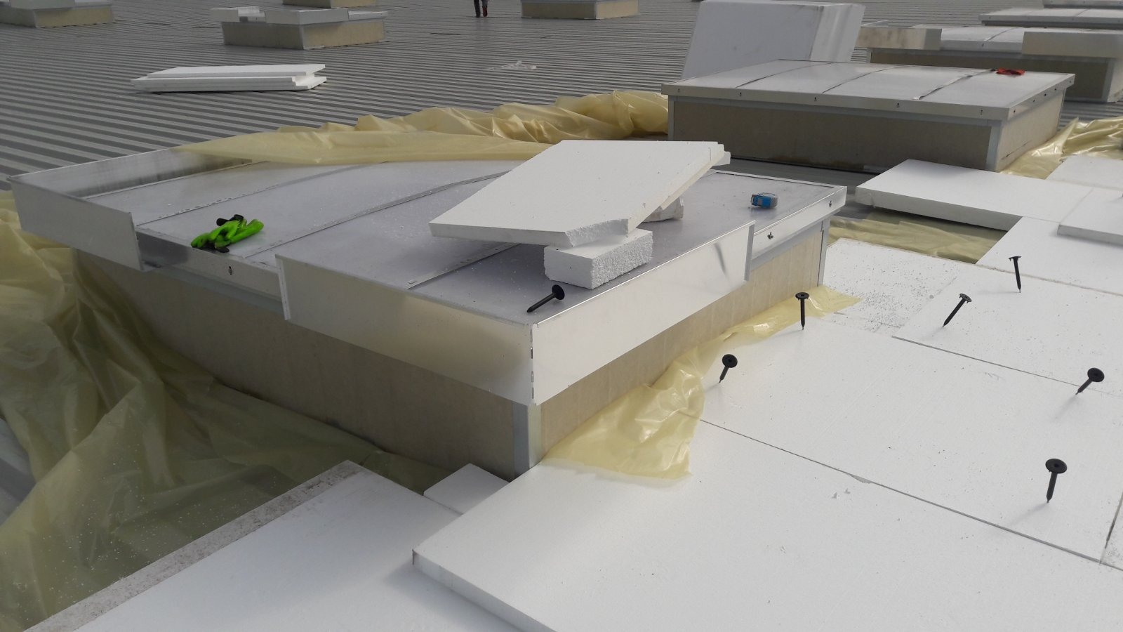 Montaż izolacji na dachu płaskim z użyciem gwoździ dekarskich, widoczne elementy wentylacyjne i fragmenty folii ochronnej.