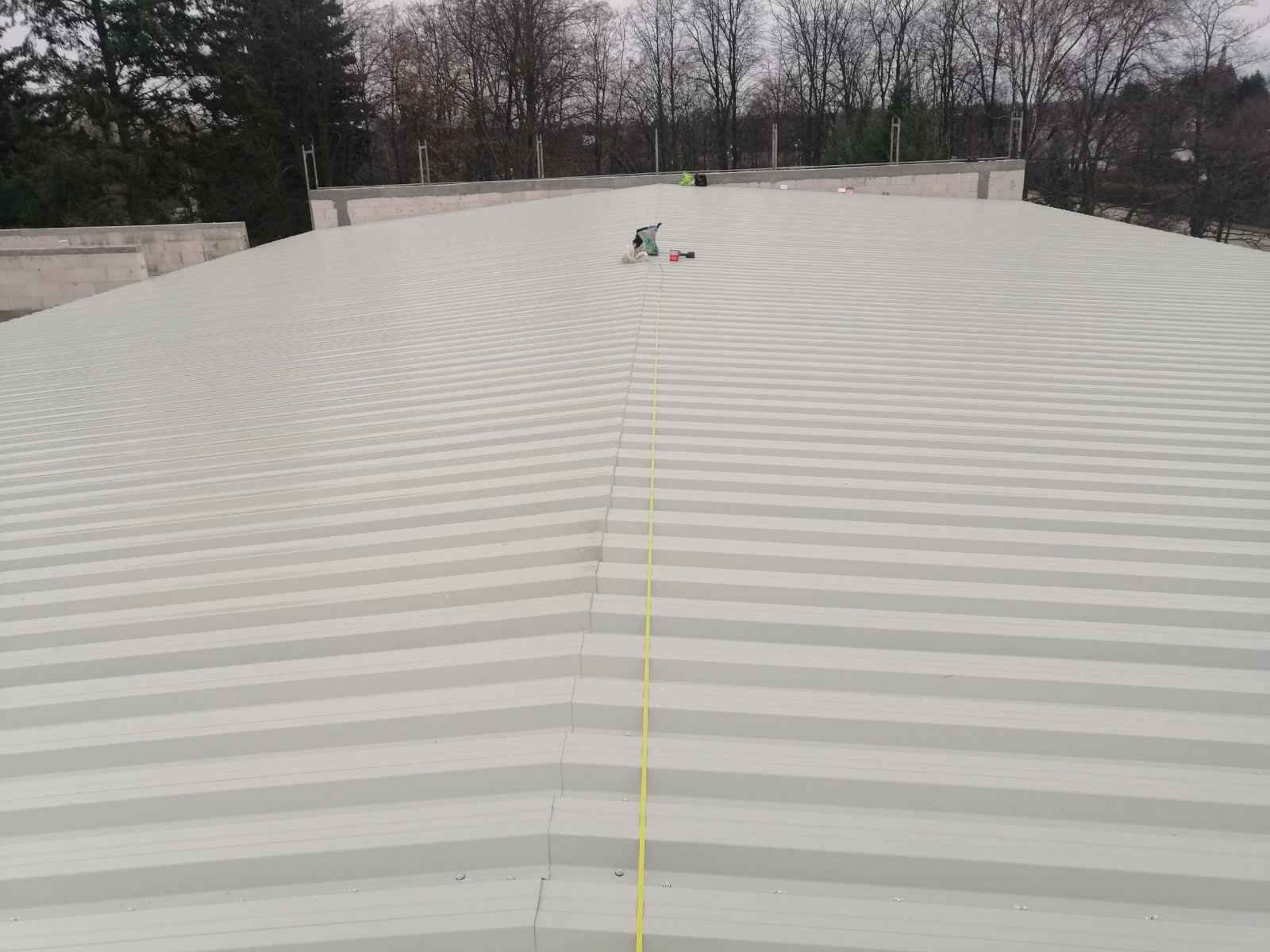 Szary dach z blachy trapezowej, widoczne narzędzia dekarskie i linka pomiarowa, tło z drzewami bez liści.