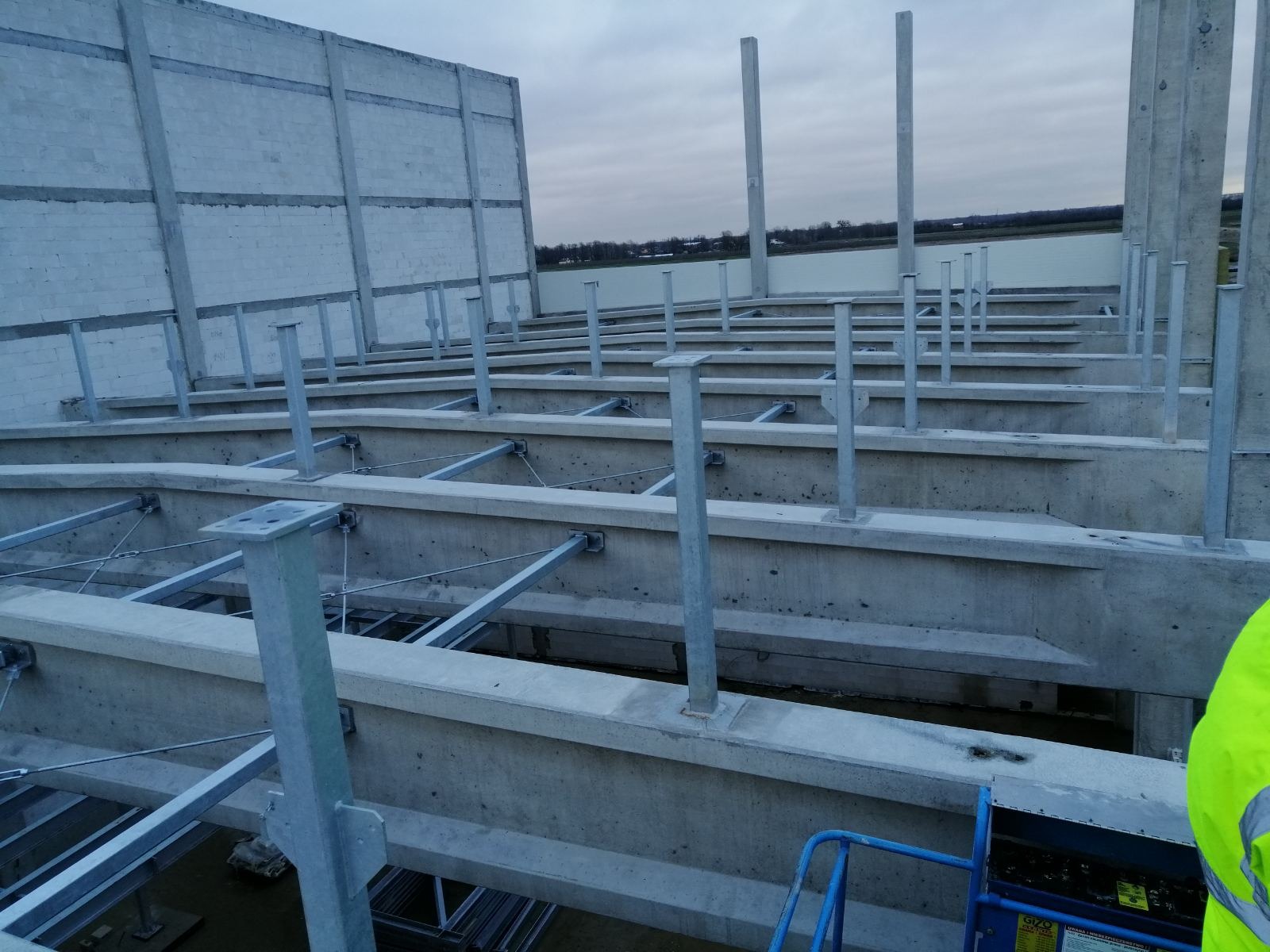 Konstrukcja dachu budynku przemysłowego z betonu i metalu, widoczne elementy nośne i wsporniki, w tle szary mur i pochmurne niebo.