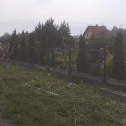 Montaż ogrodzenia panelowego z podmurówką betonową, widoczne tuje posadzone wzdłuż ogrodzenia, teren po robotach porządkowany przez osoby z grabiami.