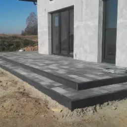 Nowoczesny taras z szarej kostki brukowej prowadzący do przeszklonych drzwi domu z surową elewacją, widoczne dwustopniowe schody i przygotowany teren wokół.