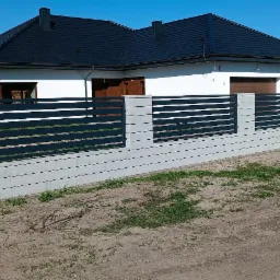 Nowoczesne ogrodzenie z szarych betonowych bloczków i poziomych, czarnych przęseł, na tle białego domu z ciemnym dachem i budowy w stanie surowym otwartym.