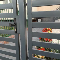 Nowoczesna, szara brama wjazdowa z poziomymi profilami, widok z bliska na klamkę i fragment ogrodu z czerwonymi i żółtymi kwiatami w tle.