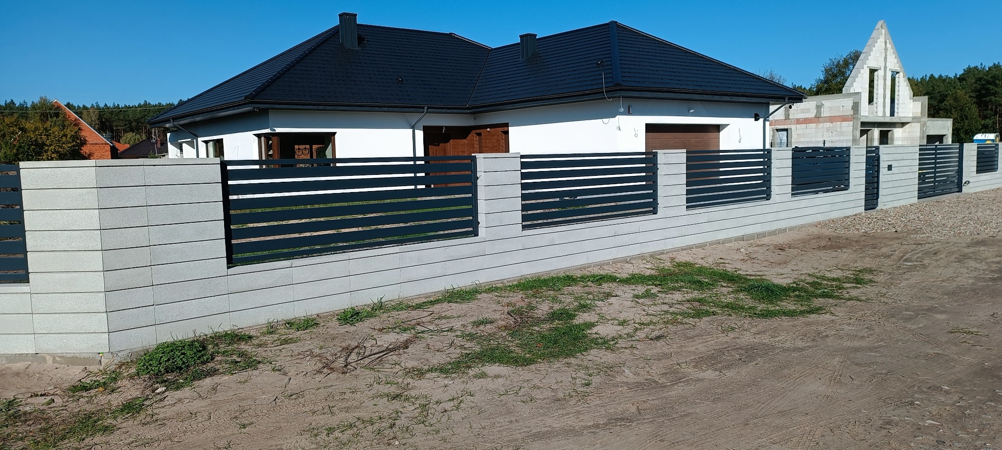 Nowoczesne ogrodzenie z szarych betonowych bloczków i poziomych, czarnych przęseł, na tle białego domu z ciemnym dachem i budowy w stanie surowym otwartym.