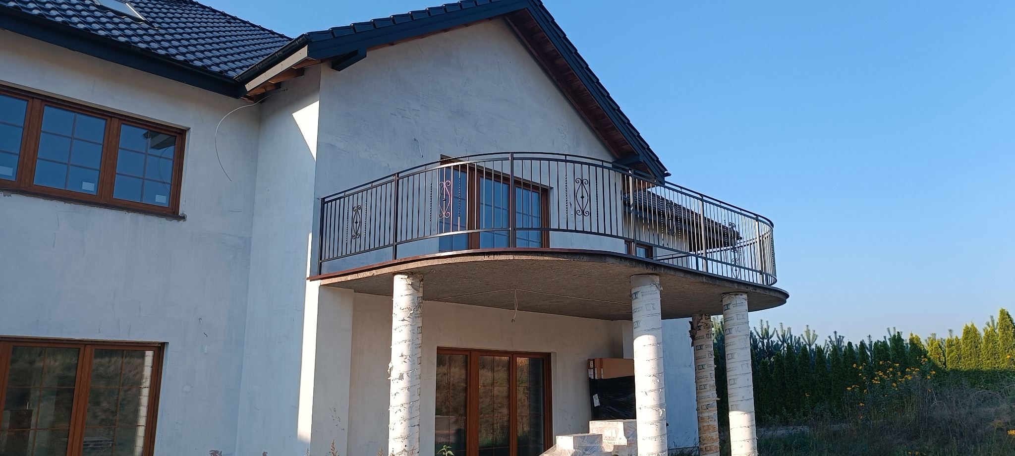 Metalowa balustrada balkonowa z ozdobnymi elementami na zaokrąglonym balkonie domu w Barcinie, widok z dołu na tle błękitnego nieba.