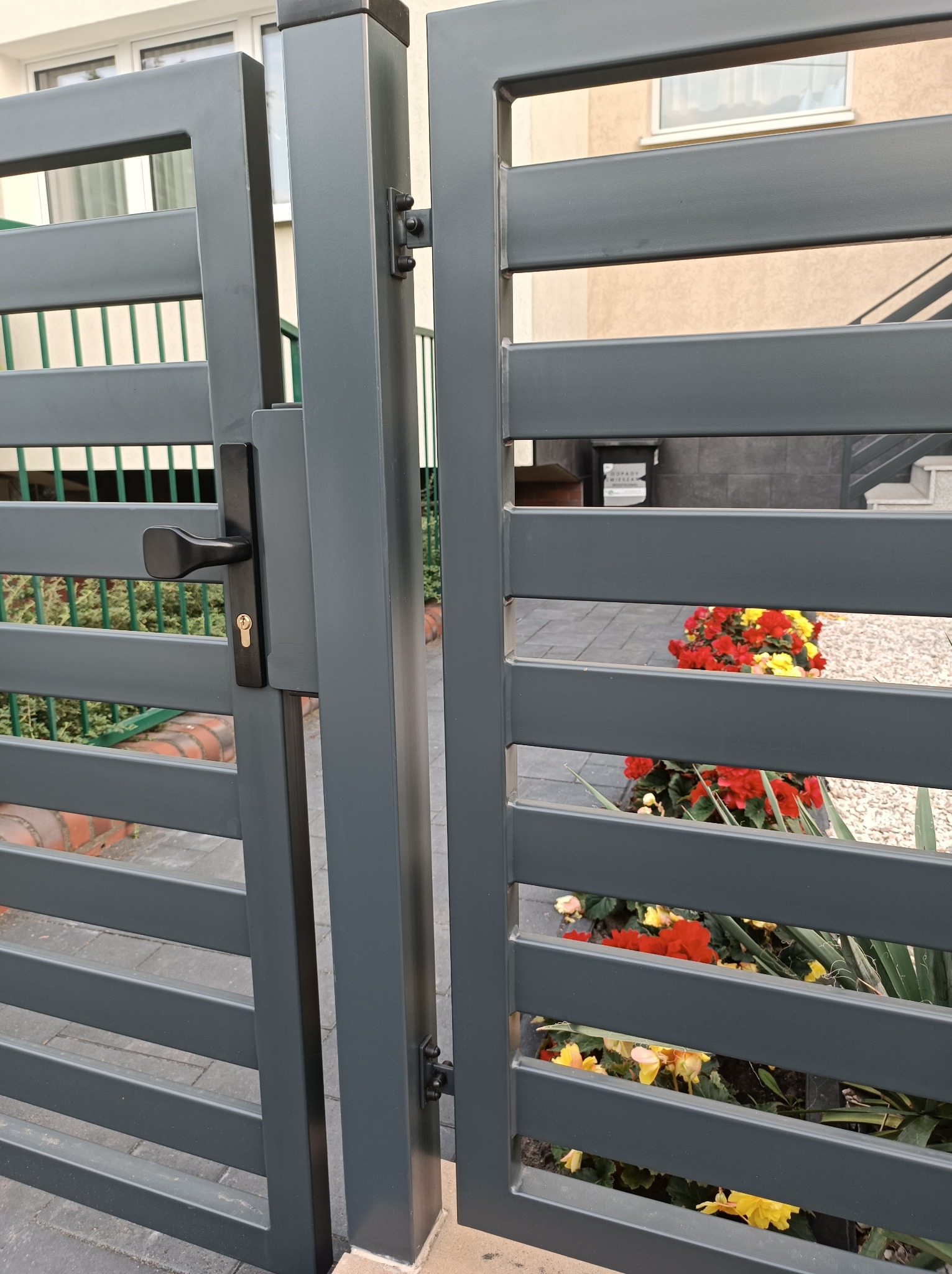 Nowoczesna, szara brama wjazdowa z poziomymi profilami, widok z bliska na klamkę i fragment ogrodu z czerwonymi i żółtymi kwiatami w tle.