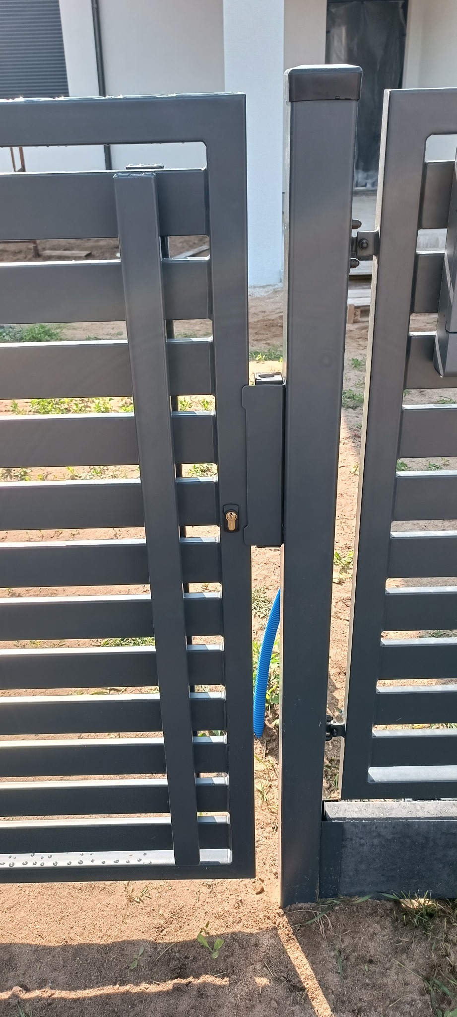 Nowoczesna, grafitowa brama wjazdowa z poziomymi szczeblami, widoczny zamek i fragment niebieskiego przewodu w ziemi. Minimalistyczny design.