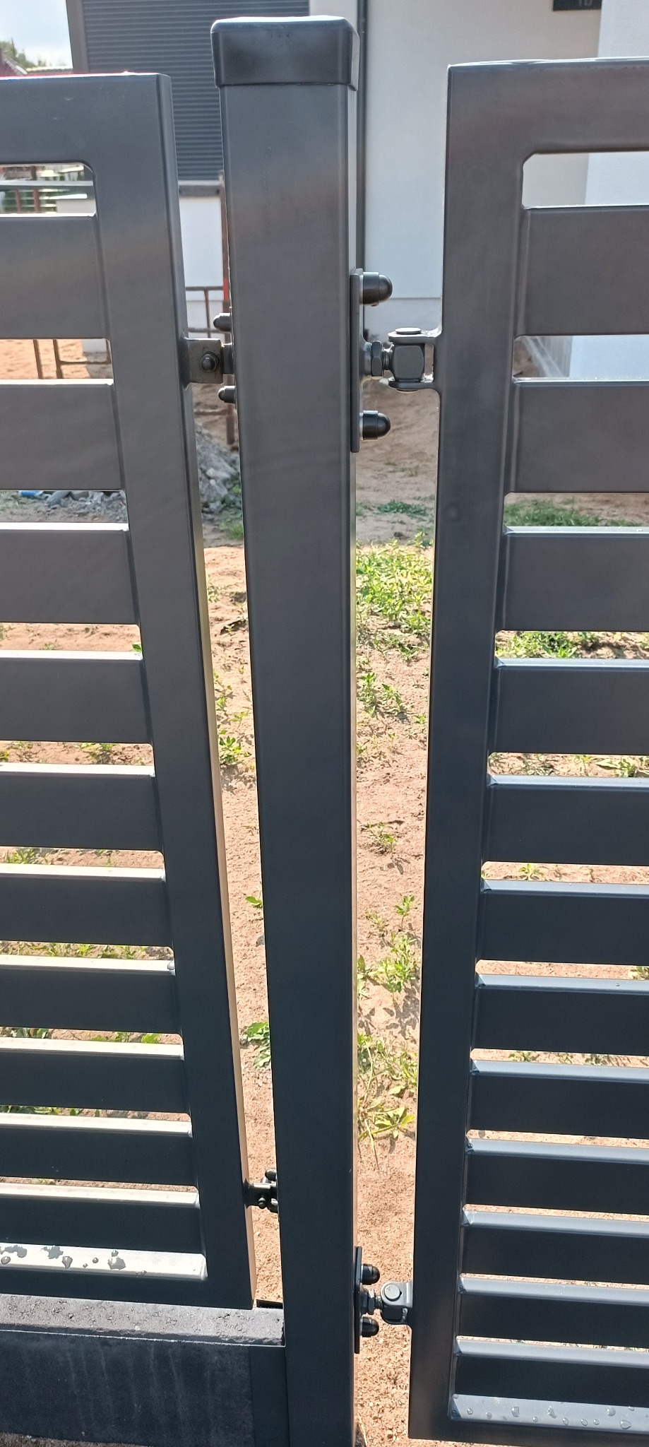 Szczegółowe ujęcie nowoczesnej, metalowej bramy wjazdowej z poziomymi szczeblami, widoczne zawiasy i elementy mocujące. Tło stanowi piaszczysta ziemia z zielenią.