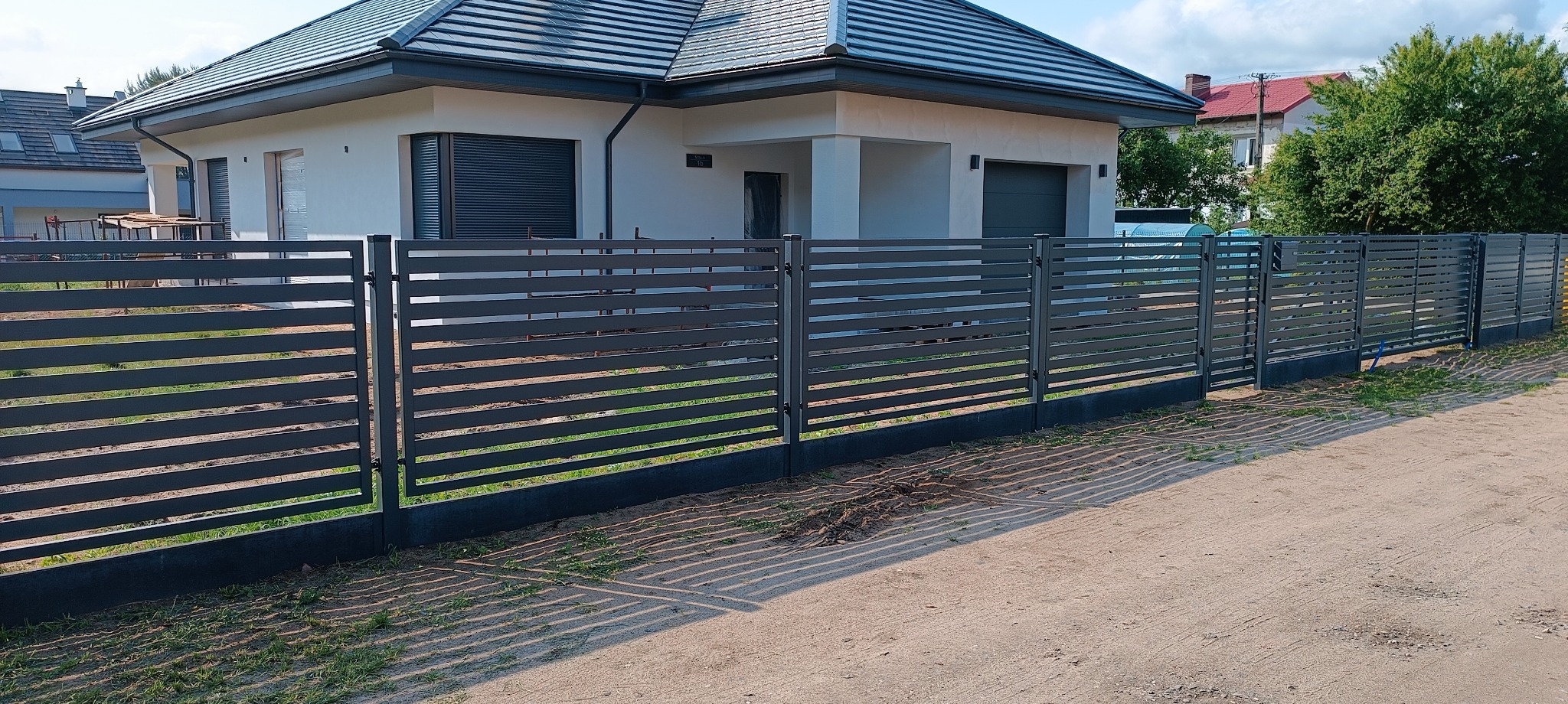 Nowoczesne, poziome ogrodzenie metalowe w kolorze grafitowym, z bramą wjazdową, na tle jasnego domu z szarą dachówką. Widoczne cienie rzucane przez ogrodzenie.