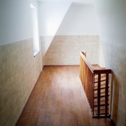 DOM - Artur Cybulski - Korytarz z drewnianą podłogą i schodami. Ściany w jasnych odcieniach beżu i bieli. Widoczne okno z lewej strony. Ujęcie z góry, perspektywa w głąb korytarza.