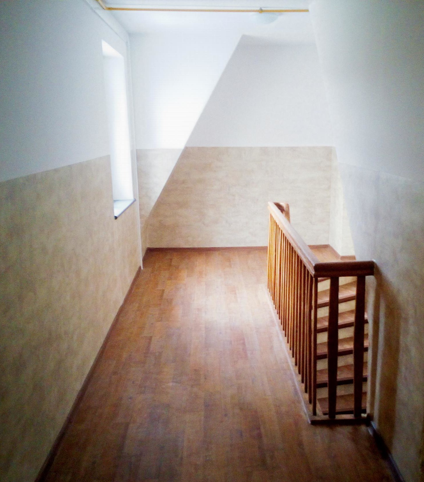 Korytarz z drewnianą podłogą i schodami. Ściany w jasnych odcieniach beżu i bieli. Widoczne okno z lewej strony. Ujęcie z góry, perspektywa w głąb korytarza.