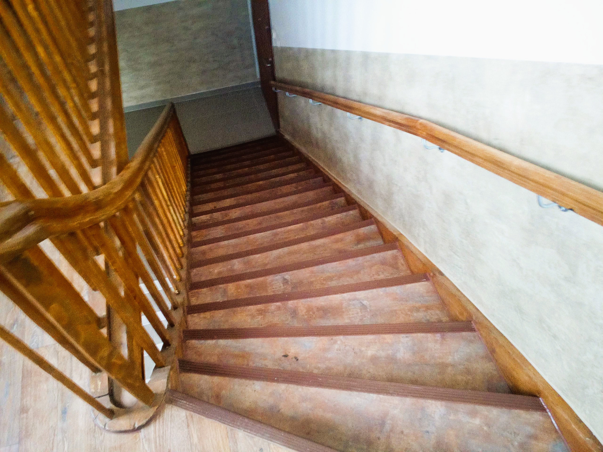 Drewniane schody z poręczą, widok z góry. Stopnie w odcieniach brązu, ściana w jasnych beżach. Charakterystyczne mocowania poręczy do ściany.